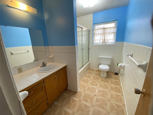 full bathroom featuring vanity, bath / shower combo with glass door, toilet, a textured ceiling, and tile walls