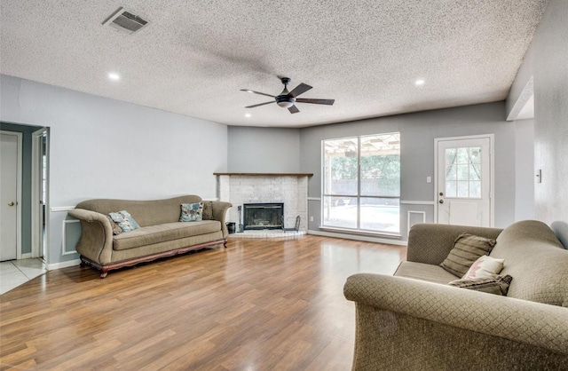 living room with ceiling fan, a textured ceiling, a brick fireplace, and light wood-type flooring