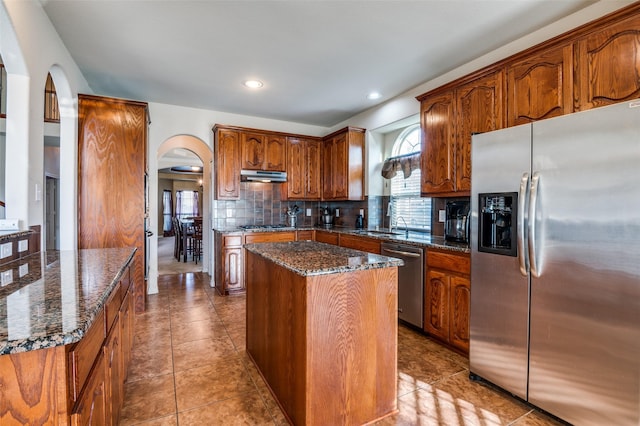 kitchen with appliances with stainless steel finishes, light tile patterned floors, dark stone counters, a kitchen island, and decorative backsplash