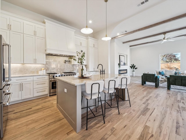 kitchen featuring white cabinetry, custom range hood, an island with sink, hanging light fixtures, and high quality appliances