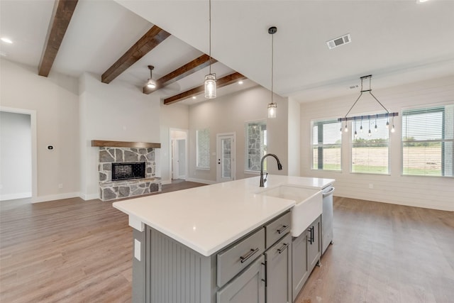 kitchen featuring gray cabinetry, a stone fireplace, ceiling fan, an island with sink, and decorative light fixtures