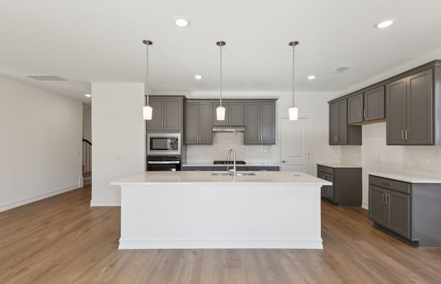 kitchen featuring decorative light fixtures, light hardwood / wood-style flooring, appliances with stainless steel finishes, gray cabinets, and an island with sink