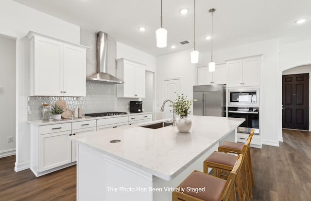 kitchen with wall chimney range hood, sink, hanging light fixtures, built in appliances, and a center island with sink