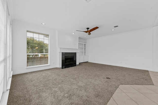 unfurnished living room featuring built in shelves, light colored carpet, ornamental molding, a tile fireplace, and ceiling fan