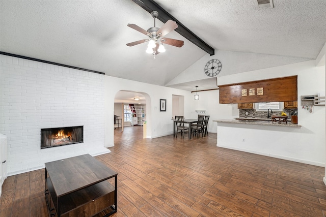 living room with ceiling fan, a brick fireplace, lofted ceiling with beams, dark hardwood / wood-style floors, and a textured ceiling