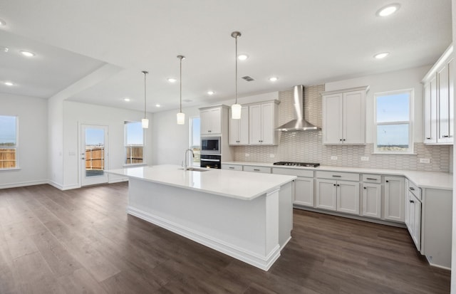 kitchen with pendant lighting, stainless steel appliances, a center island with sink, wall chimney range hood, and white cabinets