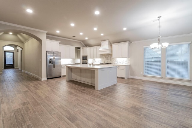 kitchen featuring appliances with stainless steel finishes, tasteful backsplash, custom exhaust hood, a kitchen island with sink, and decorative light fixtures