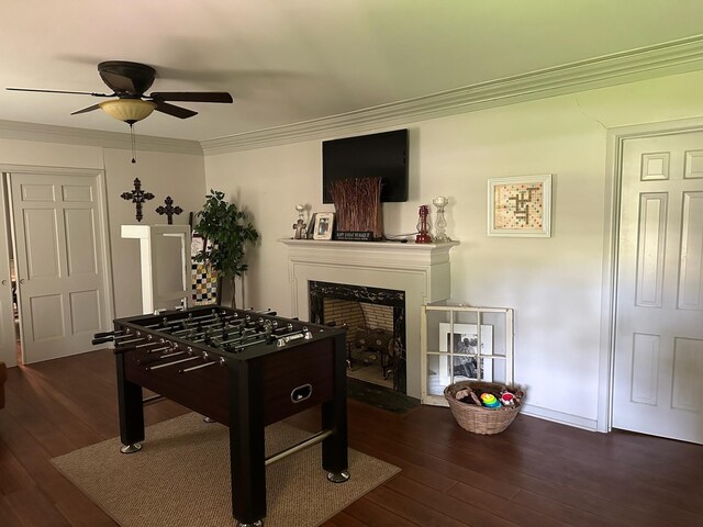 recreation room with a fireplace, ceiling fan, crown molding, and dark wood-type flooring