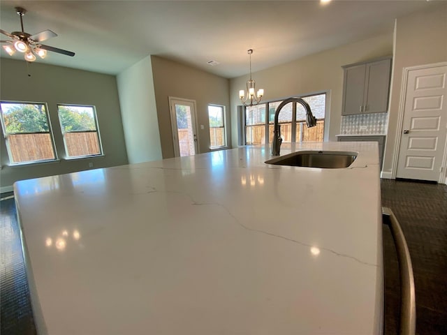 kitchen with sink, light stone counters, backsplash, decorative light fixtures, and a center island with sink