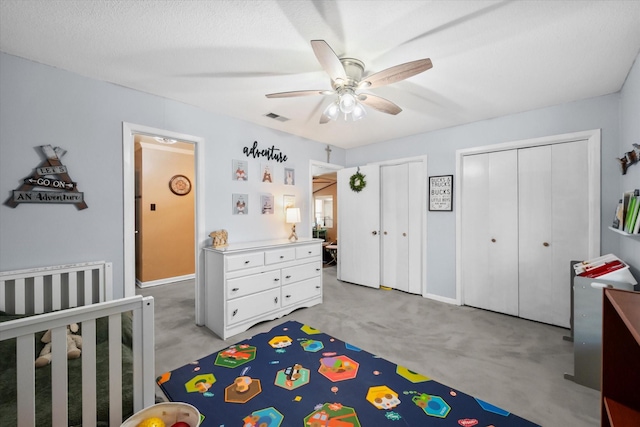 bedroom featuring ceiling fan, a textured ceiling, light carpet, a nursery area, and two closets