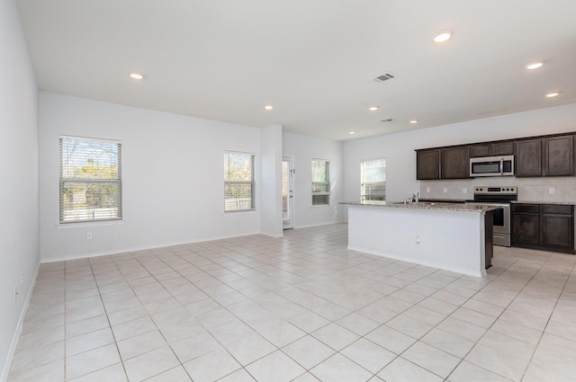 kitchen with appliances with stainless steel finishes, a kitchen island with sink, plenty of natural light, light stone countertops, and decorative backsplash