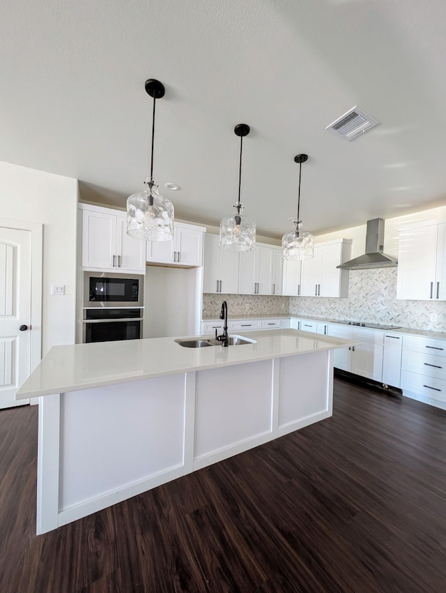 kitchen featuring built in microwave, sink, white cabinetry, an island with sink, and wall chimney range hood