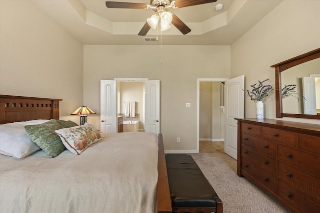 bedroom featuring a raised ceiling, ceiling fan, ensuite bathroom, and light carpet