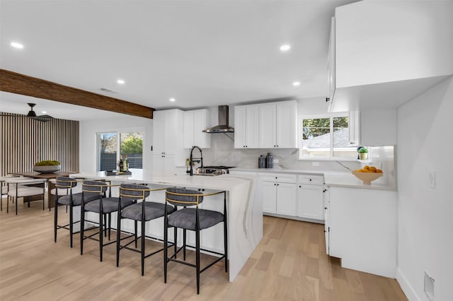 kitchen featuring white cabinets, a breakfast bar, wall chimney range hood, beamed ceiling, and light hardwood / wood-style floors