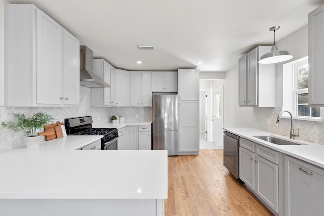 kitchen with wall chimney range hood, sink, appliances with stainless steel finishes, decorative light fixtures, and light wood-type flooring