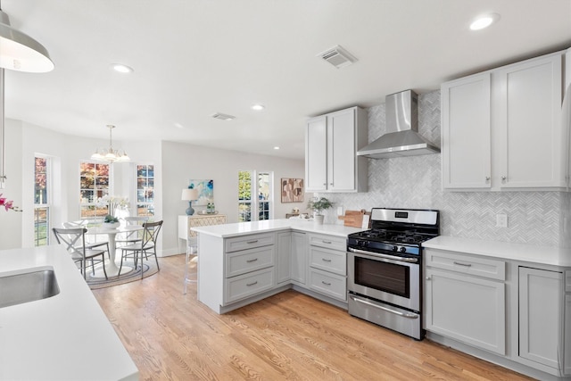 kitchen featuring kitchen peninsula, decorative backsplash, stainless steel gas range oven, wall chimney range hood, and light hardwood / wood-style flooring