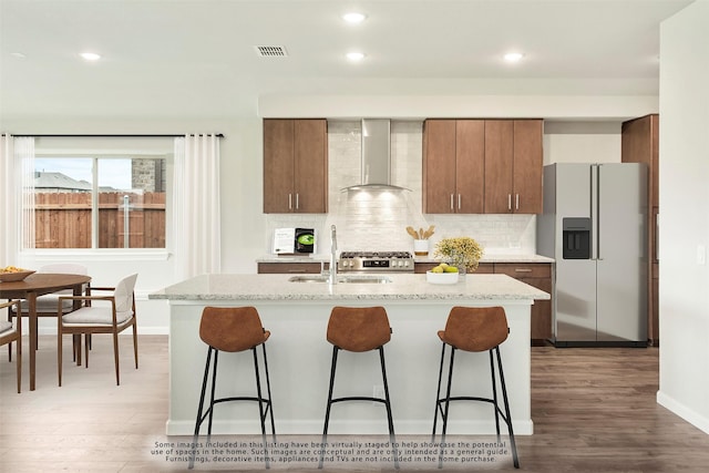 kitchen with stainless steel refrigerator with ice dispenser, wall chimney exhaust hood, sink, an island with sink, and decorative backsplash