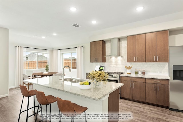 kitchen with wall chimney range hood, sink, appliances with stainless steel finishes, a kitchen breakfast bar, and a center island with sink