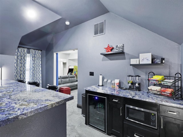 kitchen featuring stainless steel microwave, wine cooler, light stone counters, light colored carpet, and vaulted ceiling