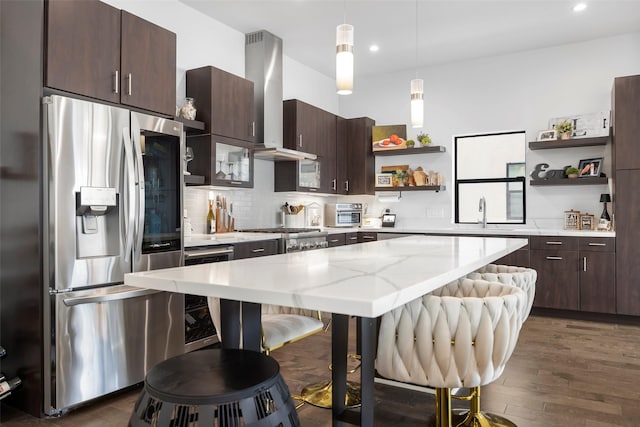 kitchen with decorative light fixtures, open shelves, stainless steel appliances, wall chimney range hood, and a kitchen bar