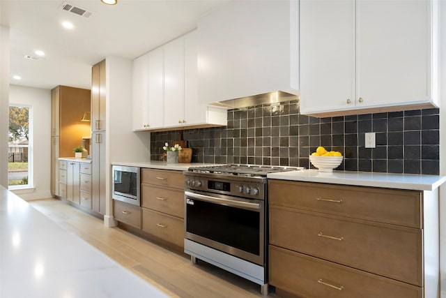 kitchen with backsplash, white cabinets, range hood, appliances with stainless steel finishes, and light hardwood / wood-style floors