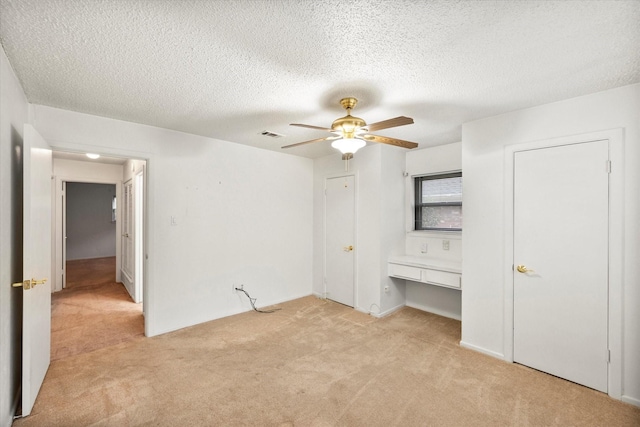 unfurnished bedroom with ceiling fan, light colored carpet, built in desk, and a textured ceiling