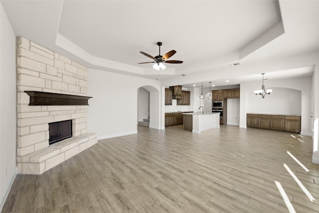 unfurnished living room featuring a stone fireplace, arched walkways, baseboards, ceiling fan with notable chandelier, and a tray ceiling
