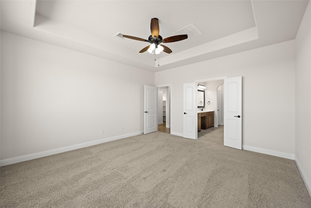 unfurnished bedroom featuring a raised ceiling, ensuite bathroom, light carpet, and ceiling fan