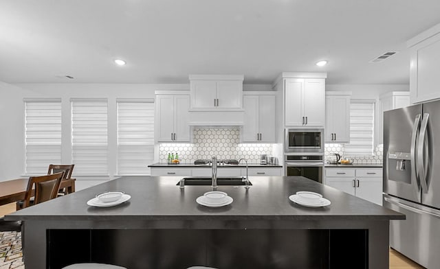 kitchen with sink, appliances with stainless steel finishes, backsplash, white cabinets, and a center island with sink