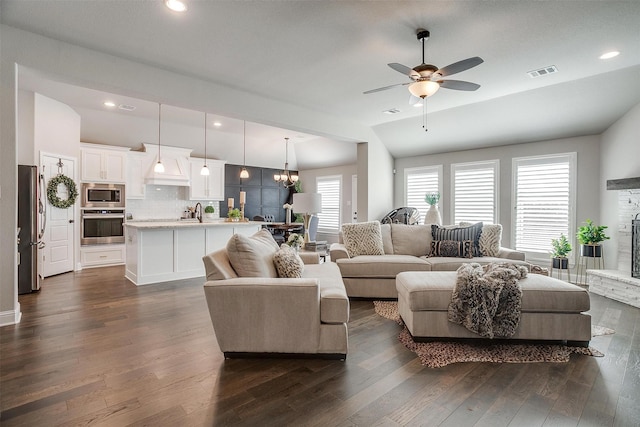 living room featuring ceiling fan with notable chandelier, vaulted ceiling, sink, dark hardwood / wood-style floors, and a stone fireplace