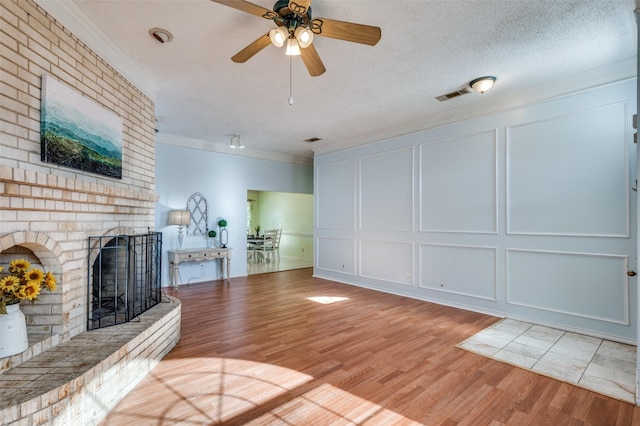 unfurnished living room featuring light wood-type flooring, a brick fireplace, a textured ceiling, ceiling fan, and crown molding