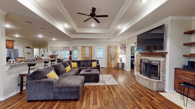 living room with ceiling fan, a brick fireplace, a raised ceiling, crown molding, and hardwood / wood-style floors