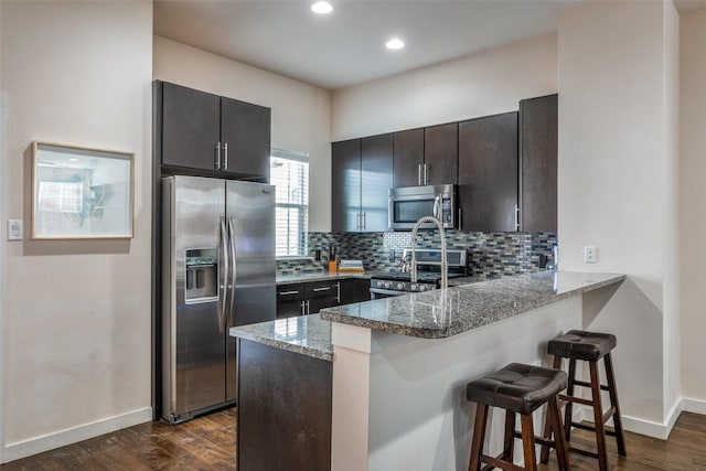 kitchen featuring a kitchen bar, dark stone countertops, appliances with stainless steel finishes, dark hardwood / wood-style flooring, and kitchen peninsula