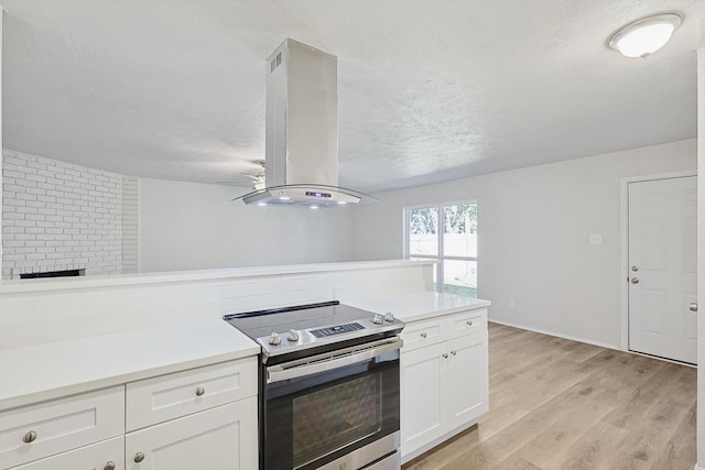 kitchen featuring island exhaust hood, a brick fireplace, white cabinetry, light hardwood / wood-style flooring, and stainless steel electric range oven