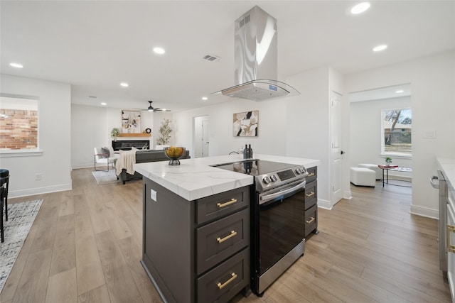 kitchen with ceiling fan, stainless steel range with electric stovetop, island range hood, a center island with sink, and light wood-type flooring