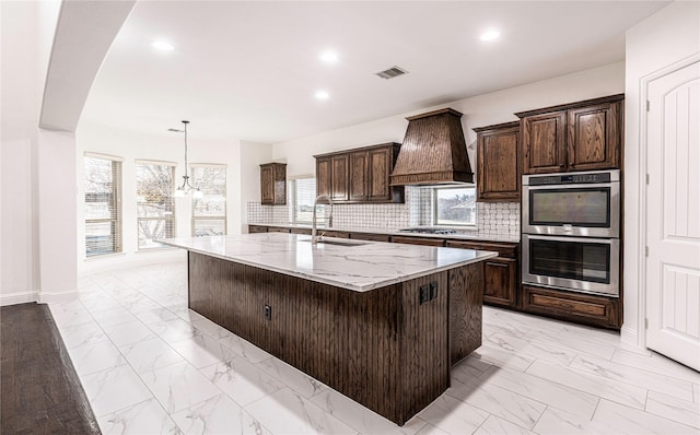 kitchen with sink, a center island with sink, appliances with stainless steel finishes, custom range hood, and pendant lighting