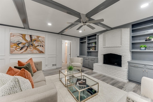 living room with a brick fireplace, built in features, beamed ceiling, ceiling fan, and light hardwood / wood-style floors