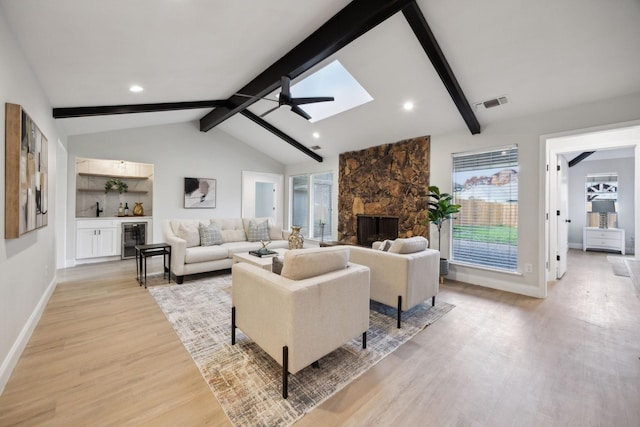 living room with ceiling fan, beverage cooler, vaulted ceiling with beams, a stone fireplace, and light hardwood / wood-style flooring