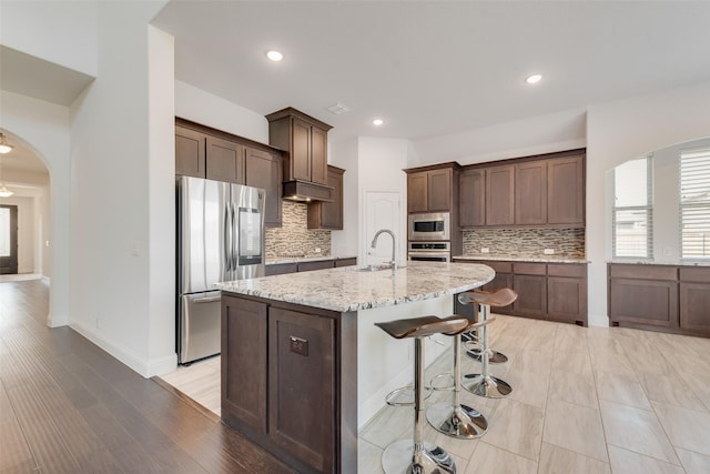 kitchen with decorative backsplash, light stone countertops, light wood-type flooring, an island with sink, and appliances with stainless steel finishes