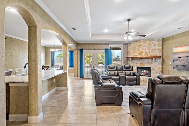 tiled living room featuring french doors, a tray ceiling, a fireplace, ceiling fan with notable chandelier, and ornamental molding