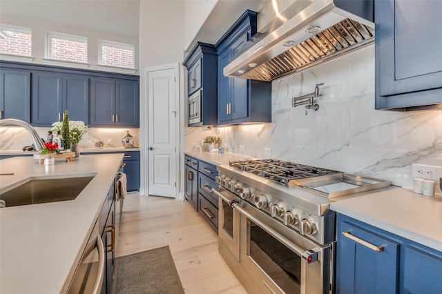 kitchen featuring wall chimney range hood, sink, stainless steel appliances, blue cabinets, and light wood-type flooring