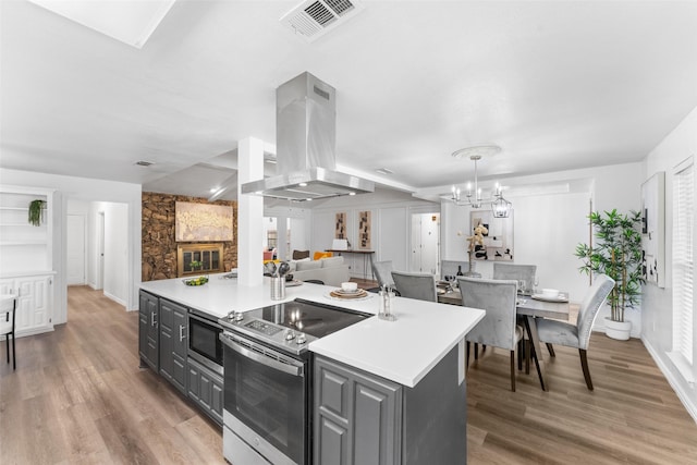 kitchen featuring light wood-type flooring, gray cabinetry, stainless steel appliances, wall chimney range hood, and an island with sink