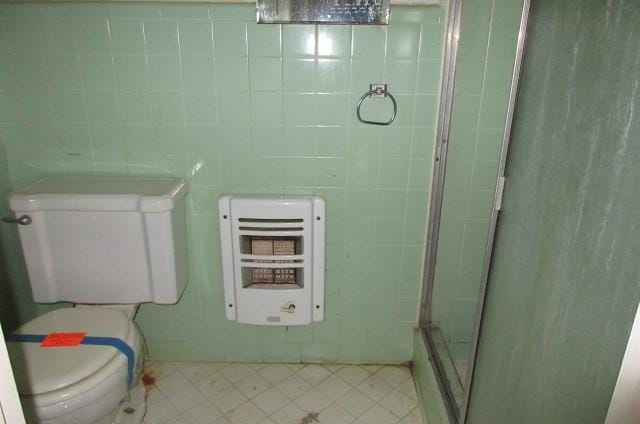 bathroom featuring a shower with door, tile walls, heating unit, tile patterned floors, and toilet