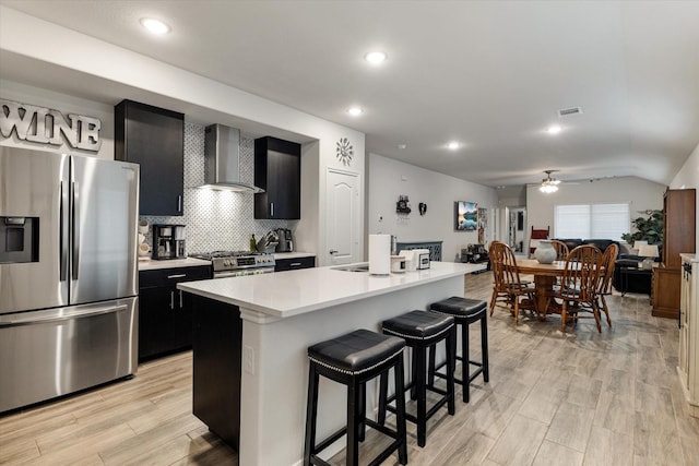 kitchen with ceiling fan, wall chimney exhaust hood, a kitchen breakfast bar, a kitchen island with sink, and appliances with stainless steel finishes