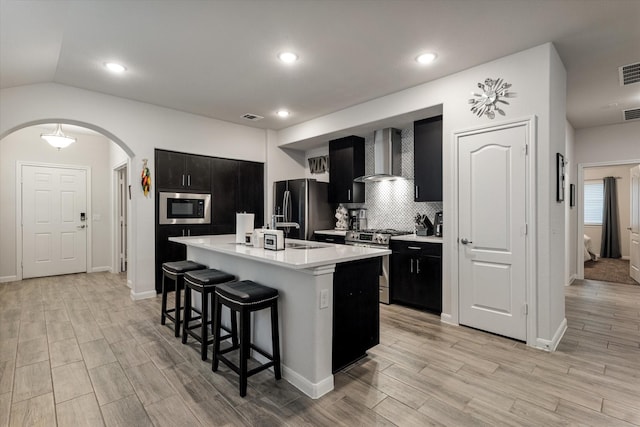 kitchen with a center island with sink, wall chimney range hood, light hardwood / wood-style flooring, a kitchen bar, and stainless steel appliances