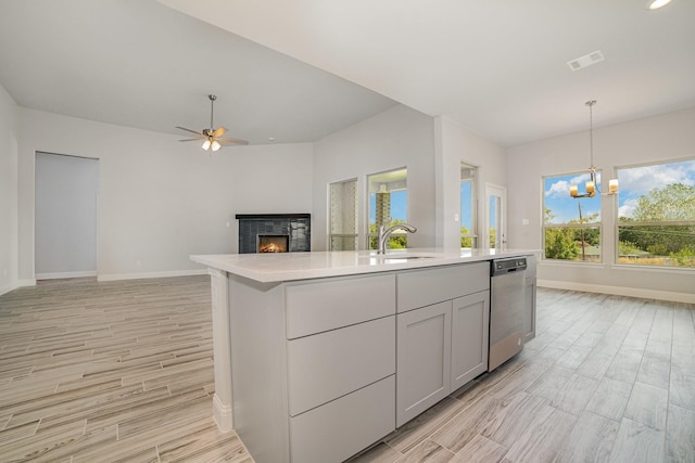 kitchen featuring stainless steel dishwasher, ceiling fan with notable chandelier, an island with sink, and light hardwood / wood-style flooring