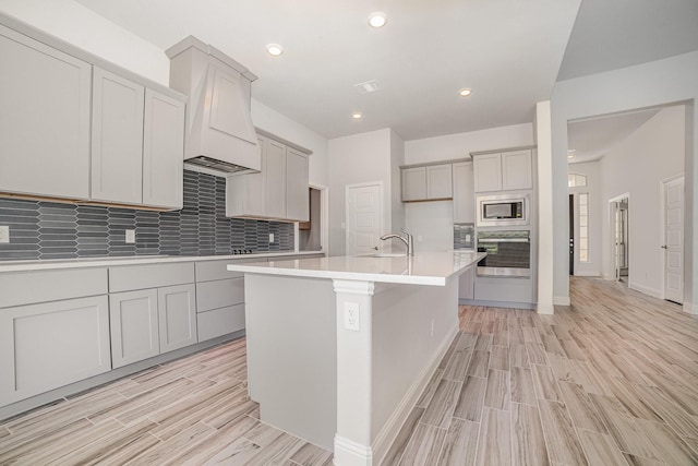 kitchen featuring decorative backsplash, light wood-type flooring, stainless steel appliances, a kitchen island with sink, and gray cabinets