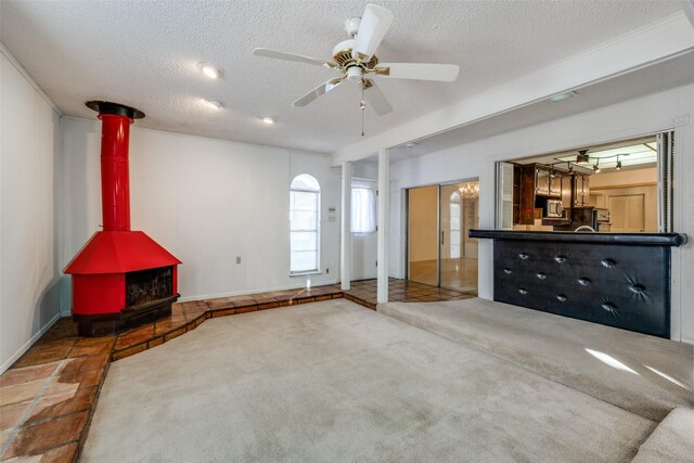 unfurnished living room with ceiling fan, a wood stove, a textured ceiling, and carpet floors