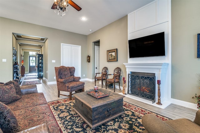 living area featuring baseboards, a ceiling fan, a warm lit fireplace, and light wood-style flooring