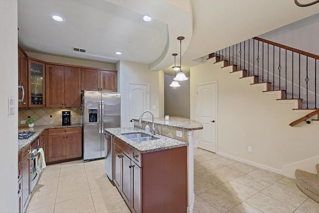 kitchen with sink, a center island with sink, appliances with stainless steel finishes, pendant lighting, and light stone countertops